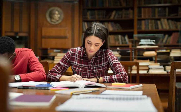 Concentrated young woman student studying in library. Female student writing notes from book. Focused girl studying and preparing an exam while sitting in public library with other university students